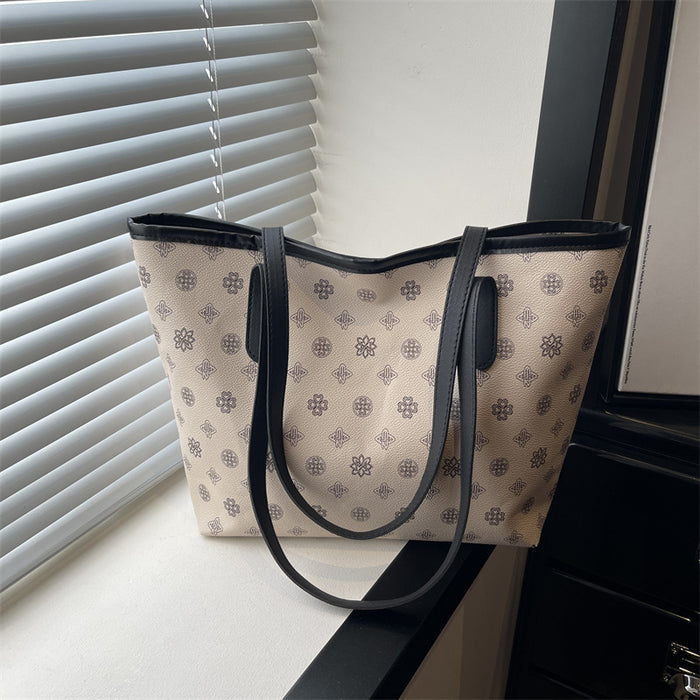 A beige tote bag with a repeating dark monogram pattern and black leather handles, resting on a windowsill next to window blinds.
