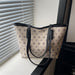 A beige tote bag with a repeating dark monogram pattern and black leather handles, resting on a windowsill next to window blinds.