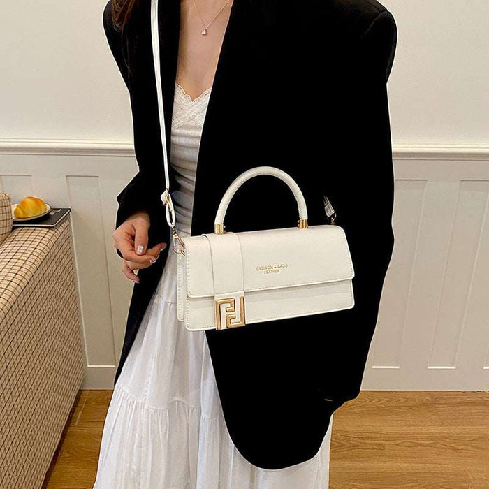 A woman wearing a black blazer and white dress carries a structured white handbag with a top handle, gold-tone hardware, and a white shoulder strap in an indoor setting.