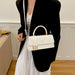 A woman wearing a black blazer and white dress carries a structured white handbag with a top handle, gold-tone hardware, and a white shoulder strap in an indoor setting.