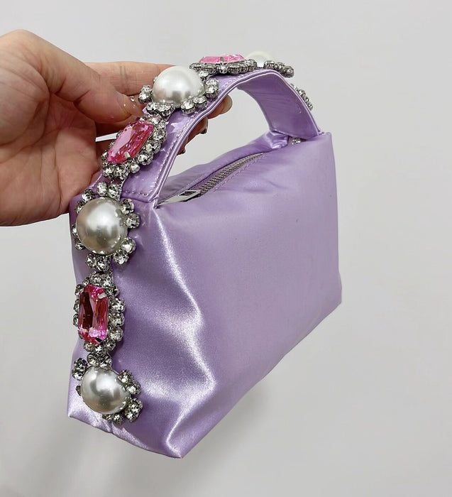 Purple handbag with decorative handle featuring pearls and pink stones, held by a hand against a neutral background.