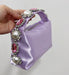 Purple handbag with decorative handle featuring pearls and pink stones, held by a hand against a neutral background.