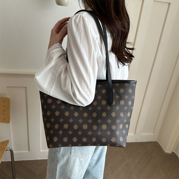 A person wearing a white long-sleeve blouse and light blue jeans carries a dark brown tote bag with a repeating gold monogram pattern and black shoulder straps in an indoor setting.