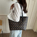 A person wearing a white long-sleeve blouse and light blue jeans carries a dark brown tote bag with a repeating gold monogram pattern and black shoulder straps in an indoor setting.