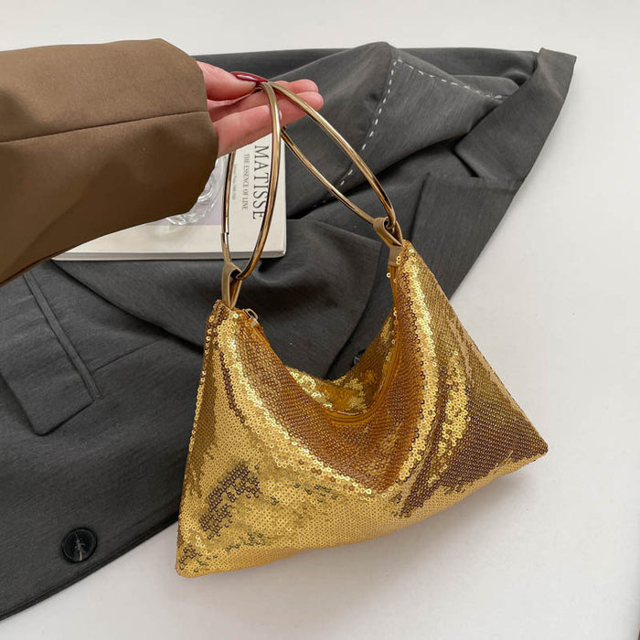A gold sequined crescent-shaped shoulder bag with thin gold handles, held by a hand wearing a brown sleeve, set against a gray blazer and white background.