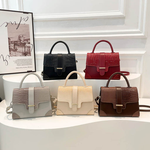 Five structured handbags with crocodile-embossed panels and top handles are displayed on white curved pedestals in an indoor showroom setting. The bags come in light gray, cream, black, burgundy red, and dark brown, each with a flap front and gold-tone hardware.