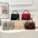 Five structured handbags with crocodile-embossed panels and top handles are displayed on white curved pedestals in an indoor showroom setting. The bags come in light gray, cream, black, burgundy red, and dark brown, each with a flap front and gold-tone hardware.