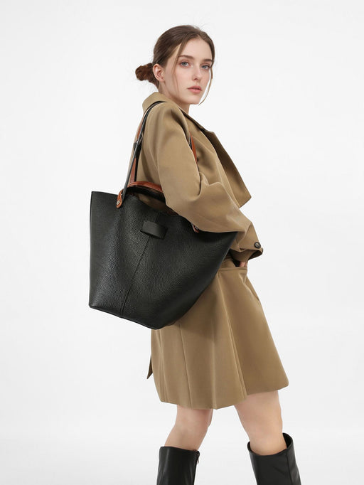A woman wearing a beige coat and black boots, carrying a large black shoulder tote bag with a lychee pattern.