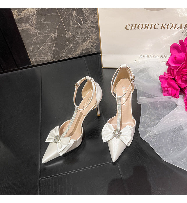 White high-heeled shoes with decorative bows on a black surface, next to a pink flower and 'Choric Koia' box.