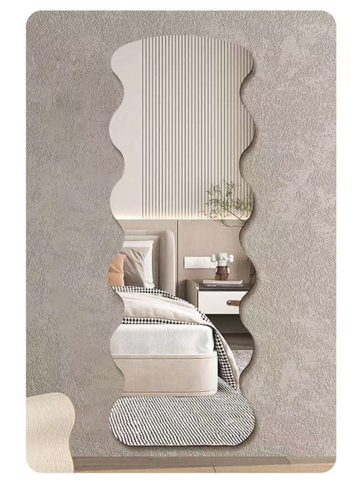 A vertically oriented wall mirror with an organic, wavy scalloped silhouette, shown mounted on a textured gray wall. The reflection in the mirror displays a modern bedroom interior with a bed, bedside table, and a small plant.