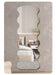 A vertically oriented wall mirror with an organic, wavy scalloped silhouette, shown mounted on a textured gray wall. The reflection in the mirror displays a modern bedroom interior with a bed, bedside table, and a small plant.