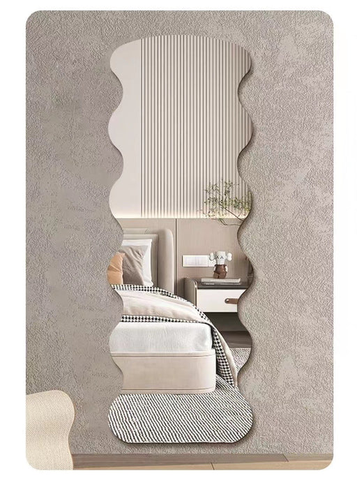 A vertically oriented wall mirror with an organic, wavy scalloped silhouette, shown mounted on a textured gray wall. The reflection in the mirror displays a modern bedroom interior with a bed, bedside table, and a small plant.