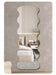 A vertically oriented wall mirror with an organic, wavy scalloped silhouette, shown mounted on a textured gray wall. The reflection in the mirror displays a modern bedroom interior with a bed, bedside table, and a small plant.