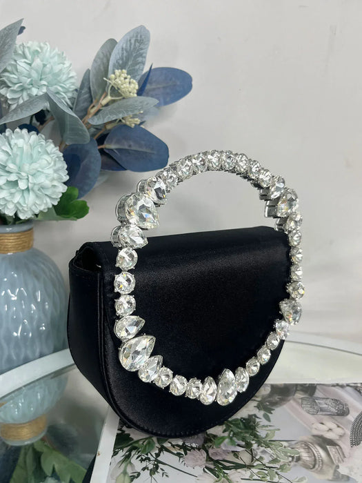 Black handbag with a decorative handle featuring crystals, placed on a surface with floral decorations.