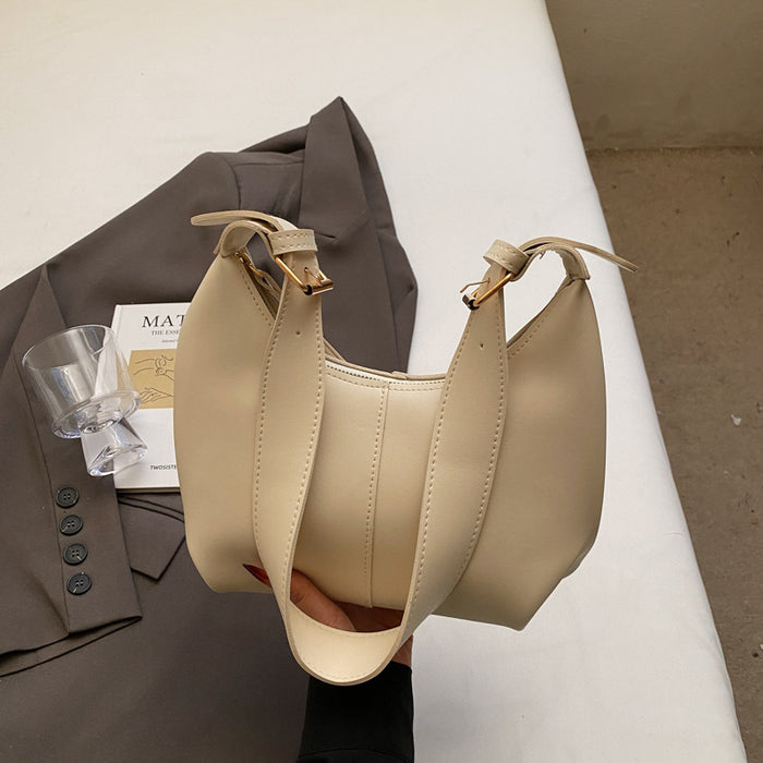 A beige faux leather slouchy shoulder bag held by a hand, styled with a brown blazer, a clear glass, and a book on a white surface.