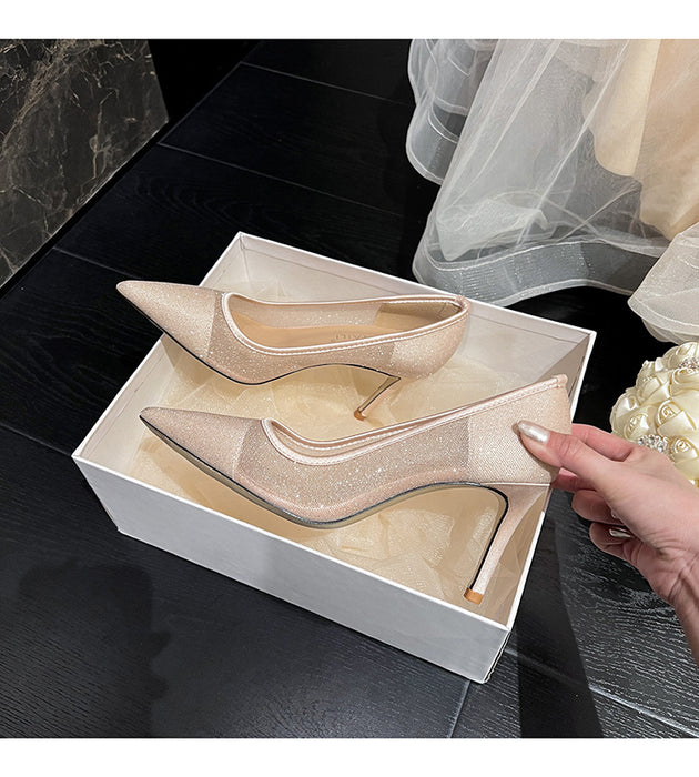 Beige high-heeled shoes in a box with a hand opening it, on a dark tiled floor.