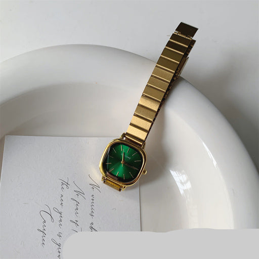 A gold-tone metal watch with a green dial and bezel, resting on a white ceramic dish over a white card with cursive writing.