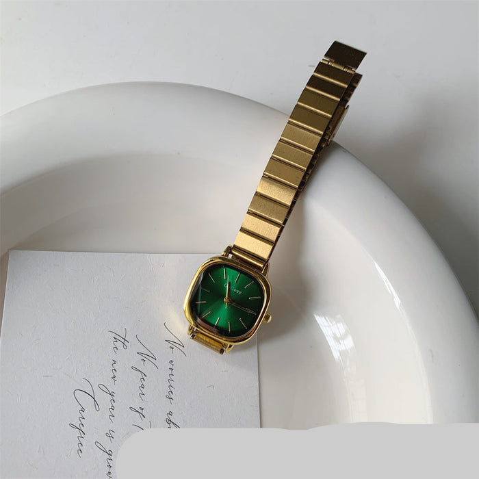 A gold-tone metal watch with a green dial and bezel, resting on a white ceramic dish over a white card with cursive writing.