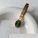 A gold-tone metal watch with a green dial and bezel, resting on a white ceramic dish over a white card with cursive writing.