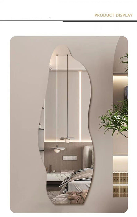 An asymmetrical, organic-shaped wall mirror with a wavy, fluid silhouette, displayed in a modern bedroom setting. The mirror is mounted on a beige wall, reflecting a bed, pendant lights, and a plant.