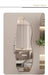 An asymmetrical, organic-shaped wall mirror with a wavy, fluid silhouette, displayed in a modern bedroom setting. The mirror is mounted on a beige wall, reflecting a bed, pendant lights, and a plant.