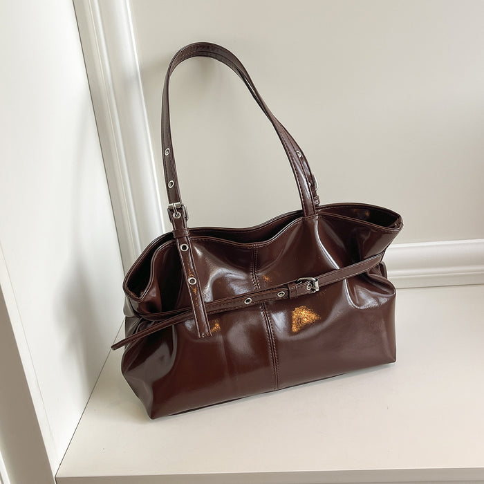 A dark brown glossy leather handbag with double shoulder straps and a front buckle strap, resting on a white shelf against a white wall.