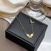 A gold necklace with a butterfly pendant and small crystal accents, displayed on a black jewelry box with draped satin fabric and a glass base in the background.