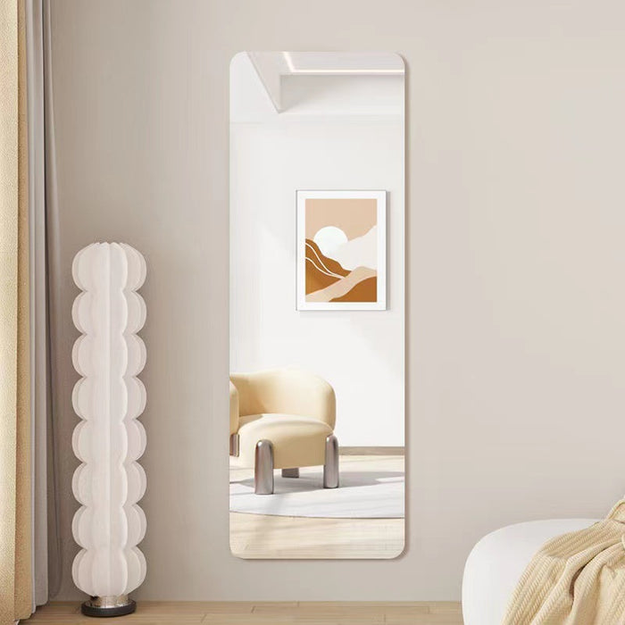 A tall, narrow rectangular wall mirror leaning against a beige wall in a modern room. The mirror reflects a cream chair and a framed abstract art print, while a white sculptural floor lamp stands to the left.