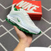 White and green sneaker held in front of a red Nike box on a tiled floor.