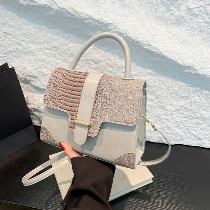 A structured handbag with a dual-tone design, featuring a textured crocodile-patterned flap in light beige and a smooth light gray body, held by a hand over a dark table against a white textured wall.