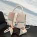 A structured handbag with a dual-tone design, featuring a textured crocodile-patterned flap in light beige and a smooth light gray body, held by a hand over a dark table against a white textured wall.