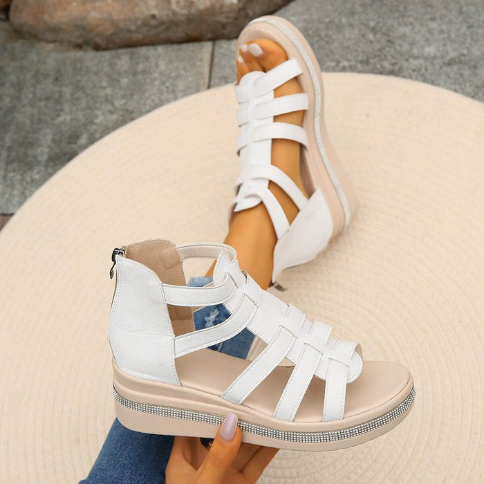 White strappy sandals with beige soles worn by a person sitting on a stone surface.
