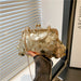 A gold metallic sequin clutch handbag with a chain strap, held by a hand wearing a dark sleeve in front of window blinds.