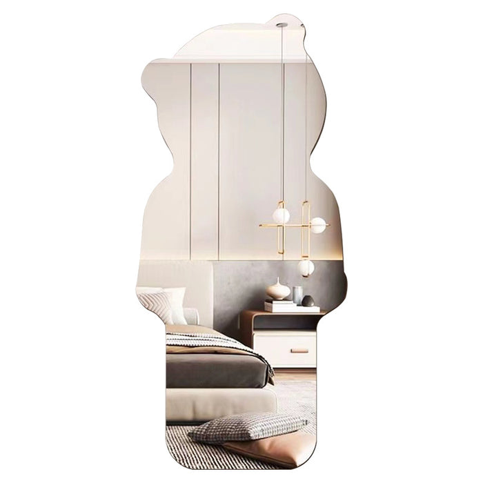 A vertically oriented decorative mirror with an organic, irregular silhouette, reflecting a modern bedroom interior with a bed, nightstand, pendant lights, and wall panels, set against a plain white background.