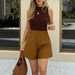 Woman wearing a brown sleeveless top and mustard yellow shorts, holding a brown handbag.