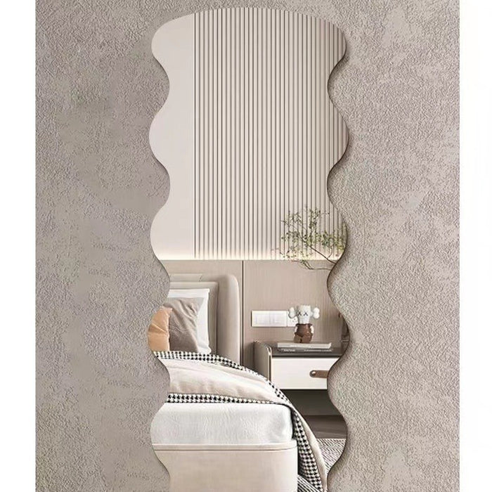 A wavy, scalloped-edge wall mirror with a textured beige frame, shown mounted on a textured gray wall and reflecting a modern bedroom interior.