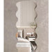 A wavy, scalloped-edge wall mirror with a textured beige frame, shown mounted on a textured gray wall and reflecting a modern bedroom interior.