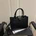 A black pebbled leather tote bag with double top handles and gold-tone hardware, displayed on a black table with a magazine underneath.