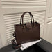 A dark brown textured leather handbag with double top handles and a detachable shoulder strap, resting on a black surface next to a white magazine in front of a white slatted door.