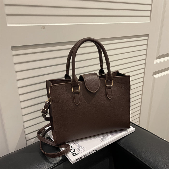 A dark brown textured leather handbag with double top handles and a detachable shoulder strap, resting on a black surface next to a white magazine in front of a white slatted door.