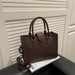 A dark brown textured leather handbag with double top handles and a detachable shoulder strap, resting on a black surface next to a white magazine in front of a white slatted door.