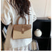 A woman from behind wearing a white blazer and carrying a structured handbag with a tan and beige woven body, brown leather top handle and flap, and a small gold clasp, set against an indoor background with a black shelf and a white vase.
