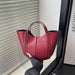 A person holds a red leather tote bag with black trim and double top handles in front of window blinds.