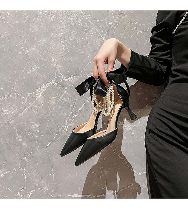 Black high-heeled shoes with bow and pearl detail held by a hand against a marble background