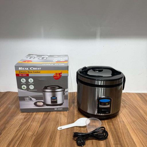 Real Crest electric rice cooker with packaging, spoon, and power cord on a wooden surface.
