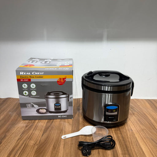 Real Crest electric rice cooker with packaging, spoon, and power cord on a wooden surface.
