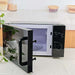 A black microwave oven with its door open, showing the interior light and glass door, placed on a light countertop with kitchen items blurred in the background.