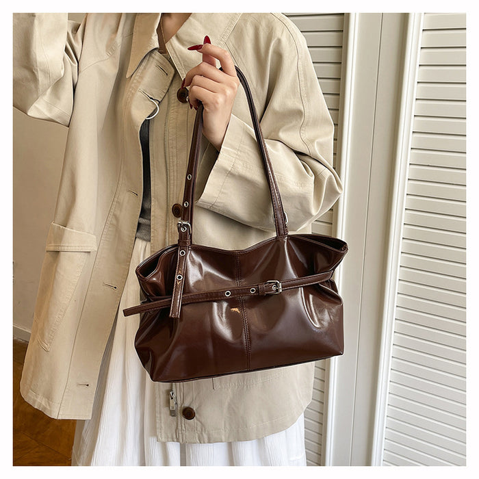 A person wearing a beige trench coat holds a dark brown leather shoulder bag with a front strap detail and silver-tone hardware, standing indoors against a white slatted door.