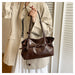 A person wearing a beige trench coat holds a dark brown leather shoulder bag with a front strap detail and silver-tone hardware, standing indoors against a white slatted door.