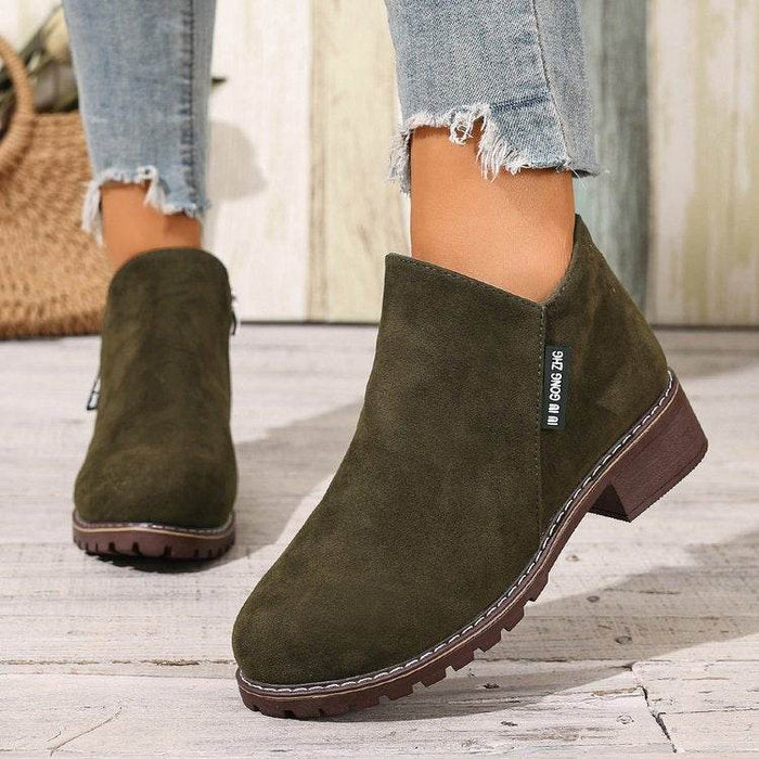Green suede ankle boots worn with denim jeans on a wooden floor.